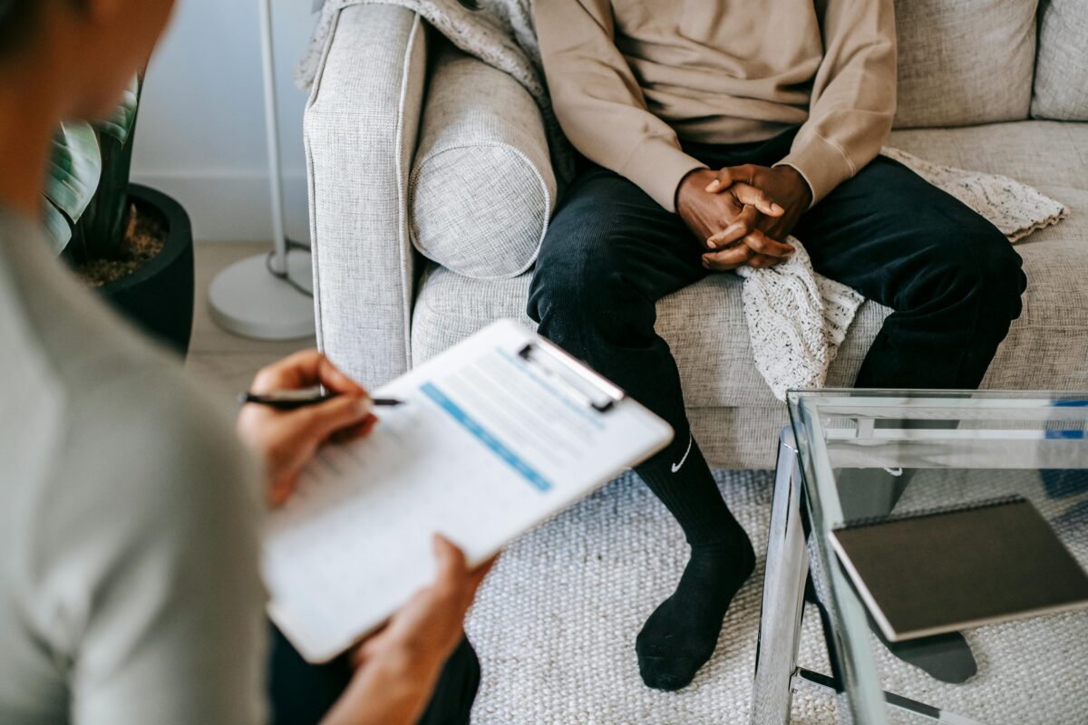 Séance de thérapie cognitivo-comportementale : un thérapeute prend des notes face à un patient assis sur un canapé, mains croisées.