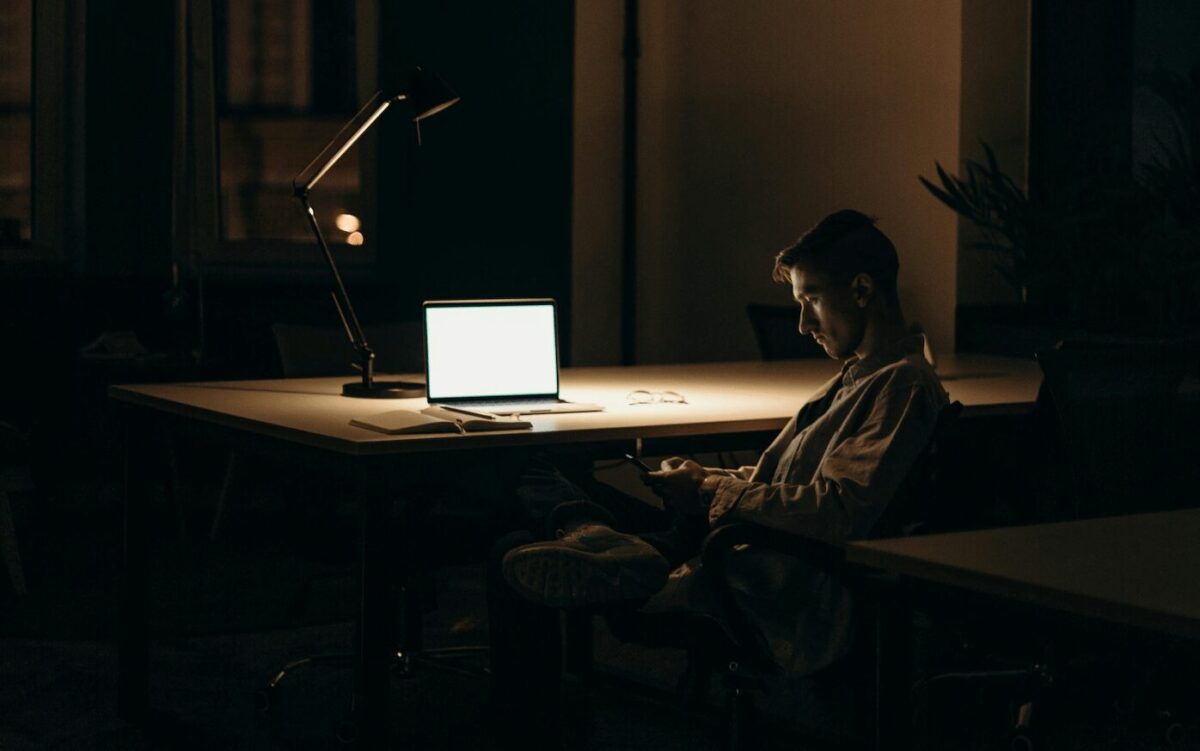 Un homme seul dans une pièce sombre, absorbé par son téléphone et éloigné de son ordinateur. Une représentation de l’attachement évitant et du repli émotionnel.