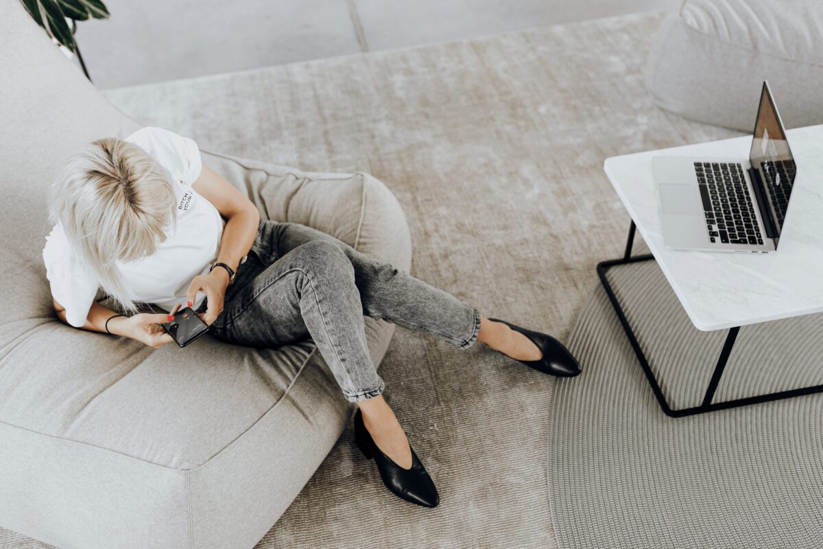 Femme allongée sur un fauteuil, absorbée par son téléphone, avec un ordinateur portable à proximité, illustrant l’addiction au téléphone et la surconnexion