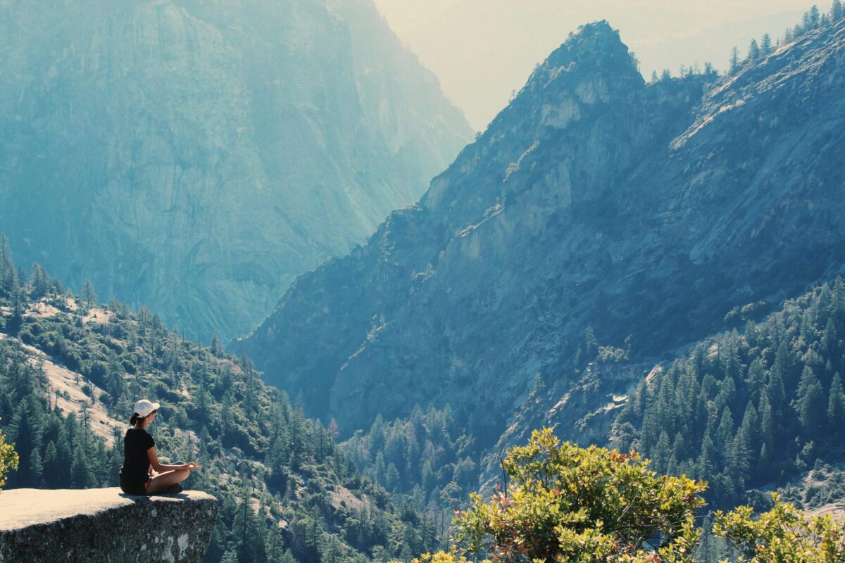Personne assise en tailleur au bord d’une falaise rocheuse, contemplant un vaste paysage de montagnes couvertes de forêt. L’atmosphère est paisible, baignée de lumière naturelle. L’image évoque la solitude, la pleine conscience et le besoin de se reconnecter à la nature, loin du tumulte quotidien.